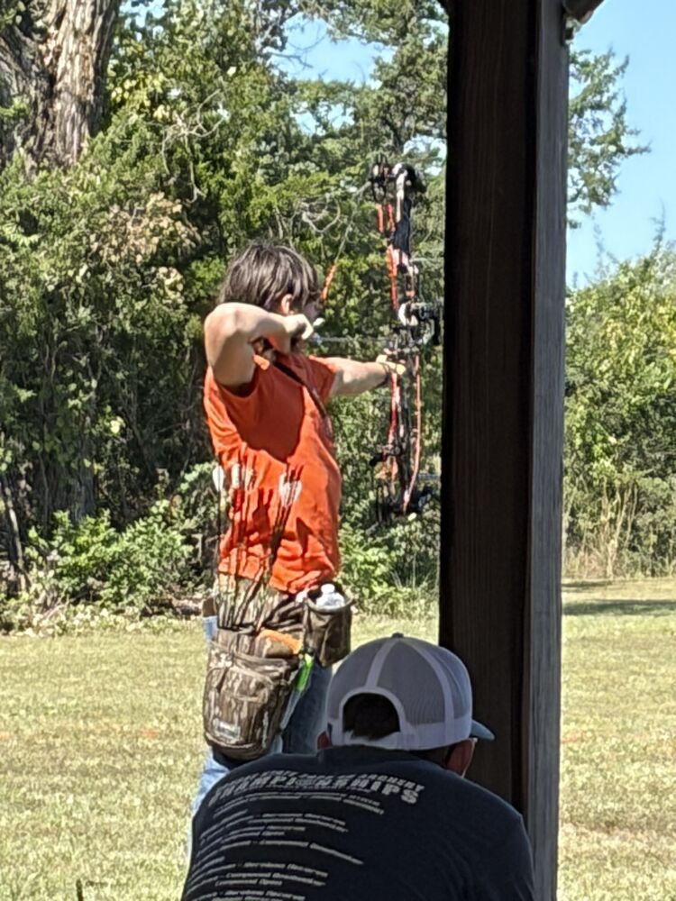 A young archer in an orange shirt at full draw with a compound bow