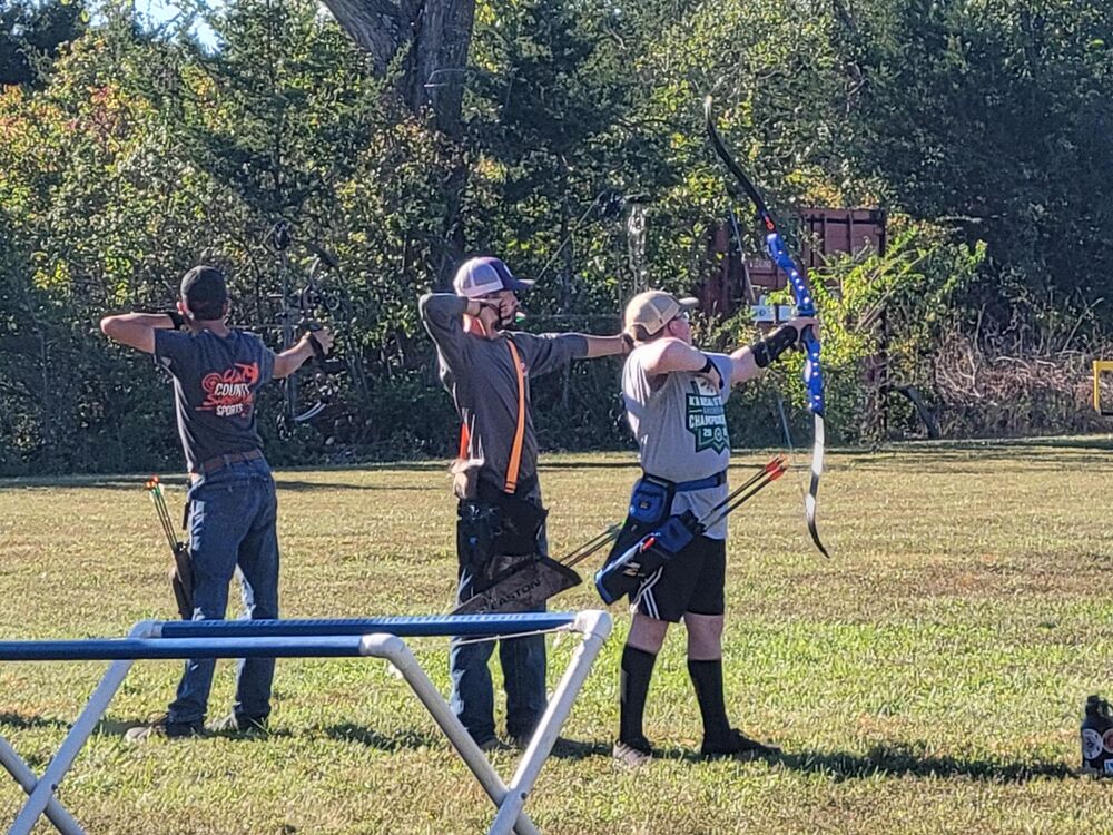 Three archers at full draw on the range, compound and recurve bows, with trees behind them