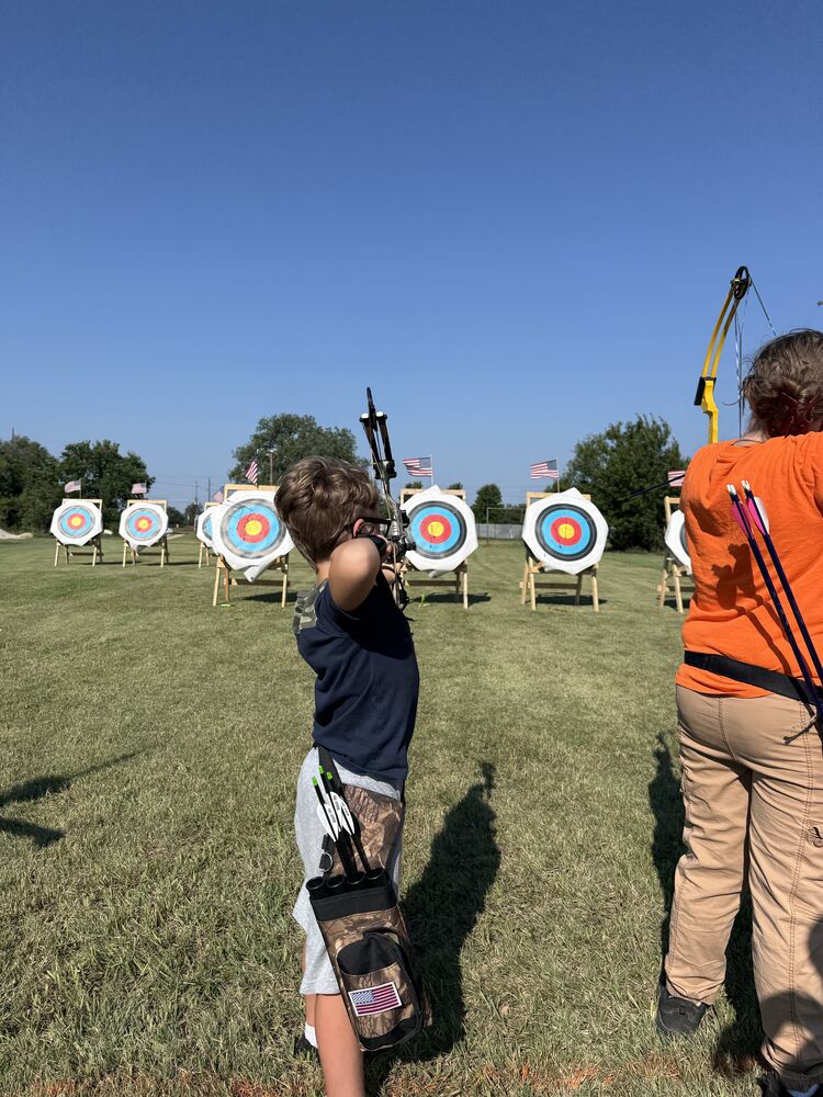 A young archer at full draw facing a row of target faces with small American flags