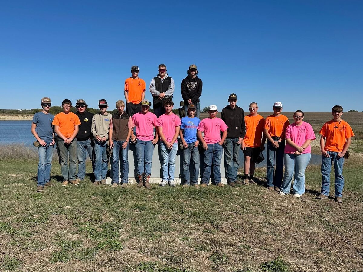 A group of trap shooters in orange and pink shirts posing together beside a pond
