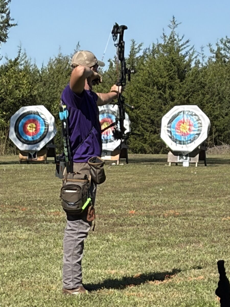 An archer in a purple shirt at full draw with a compound bow in front of target faces