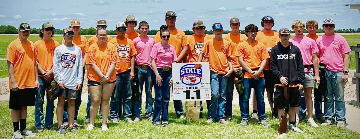 A large group of trap shooters in orange and pink shirts standing on the field behind a State tournament sign