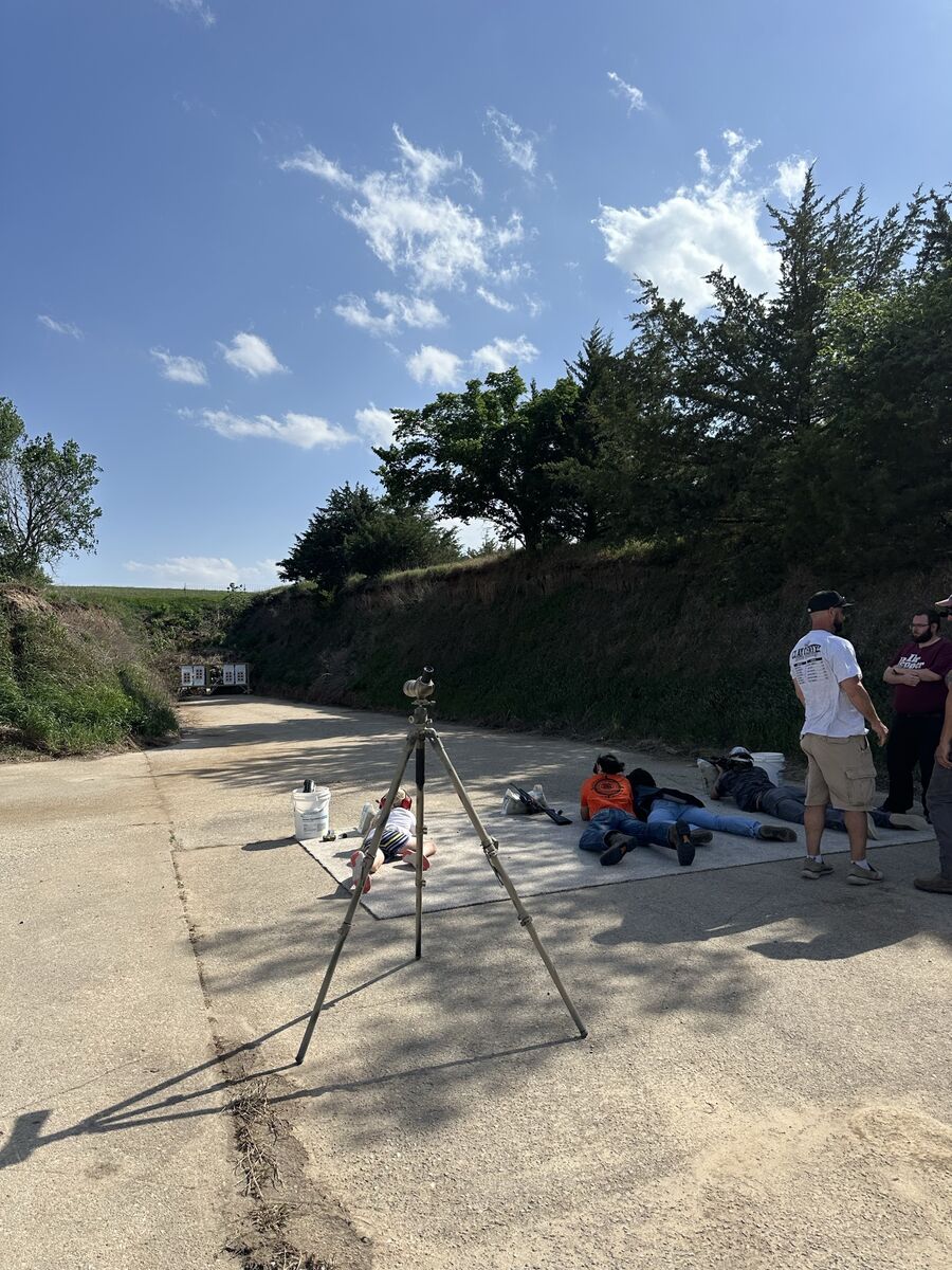 Smallbore rifle shooters lying prone on a concrete firing line with a spotting scope on a tripod while coaches stand nearby