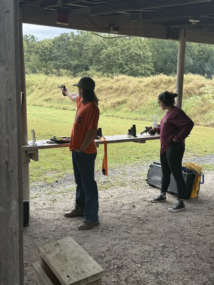 A shooter aiming a pistol downrange on an outdoor covered firing line while a second shooter handles a pistol at the bench