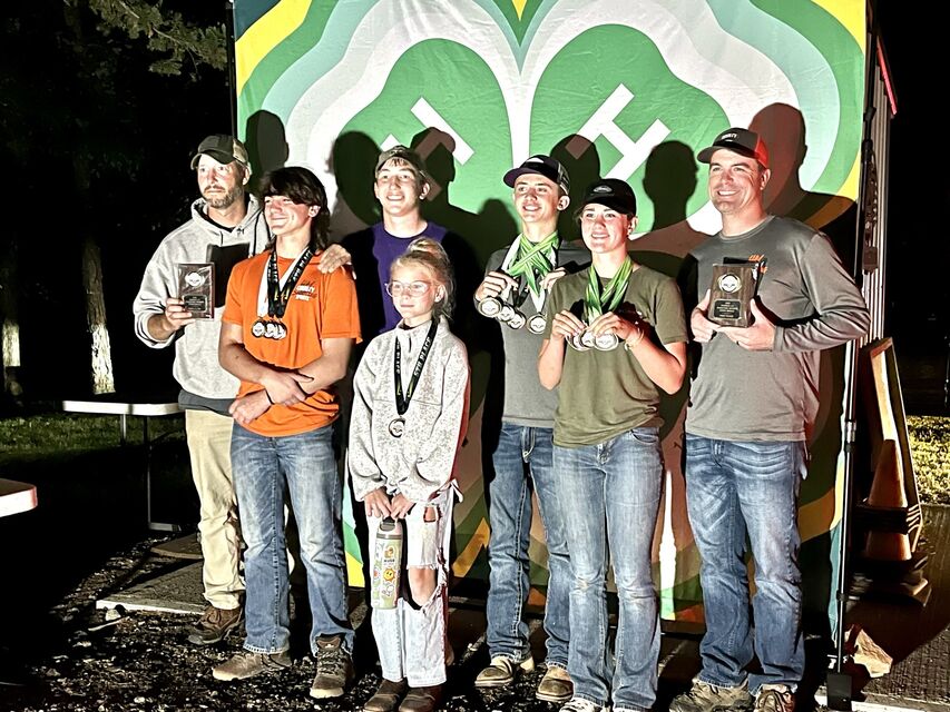 Clay County shooters and coaches holding plaques and medals in front of a 4-H clover banner at night