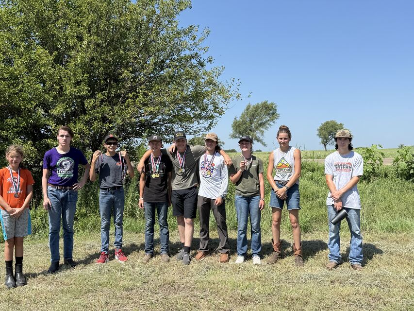 A line of ten young shooters wearing medals outdoors on a sunny day